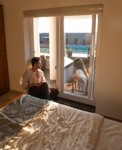 A couple relaxing in the cozy bedroom of Kappad Beach House by Kisah Stays, enjoying a warm conversation as sunlight streams through the windows. The elegant interiors, soft lighting, and serene ambiance create the perfect romantic retreat by the beach in Kozhikode, Kerala