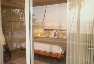 A captivating view of the room at Kappad Beach House by Kisah Stays, taken from outside the window. The glass reflects the stunning beach and Arabian Sea, blending the cozy interior with the breathtaking coastal scenery, creating a perfect harmony of luxury and nature in Kozhikode, Kerala