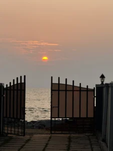 A breathtaking view of the sun peeking through the clouds above the entrance gate of Kappad Beach House by Kisah Stays. The warm golden light casts a serene glow, complementing the tranquil coastal ambiance of this beachfront retreat in Kozhikode, Kerala